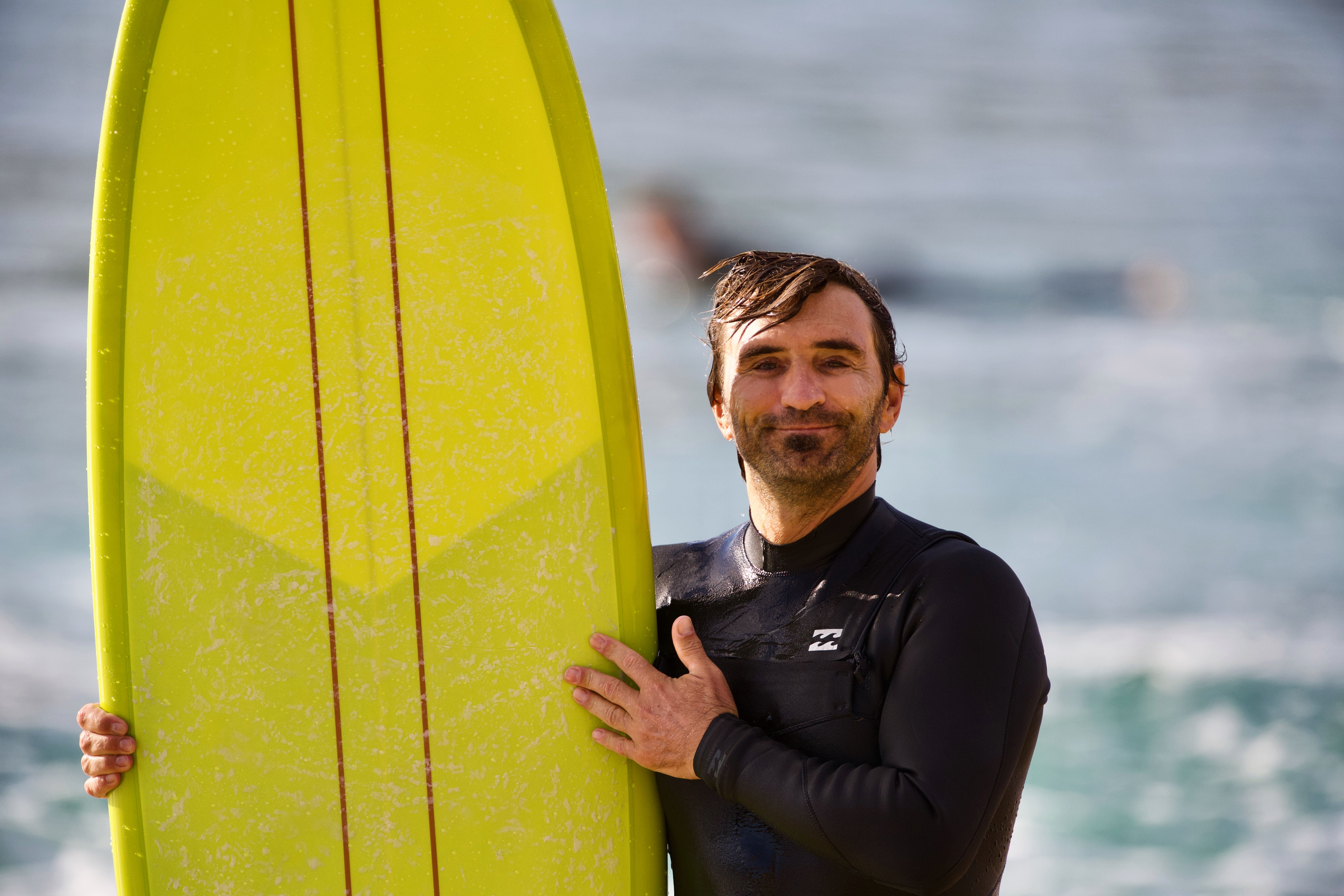 un surfeur souriant avec une combinaison de surf Billabong front zip. Il est en train de porter sa planche de surf en résine de couleur jaune dont on voit le pont avec de la wax